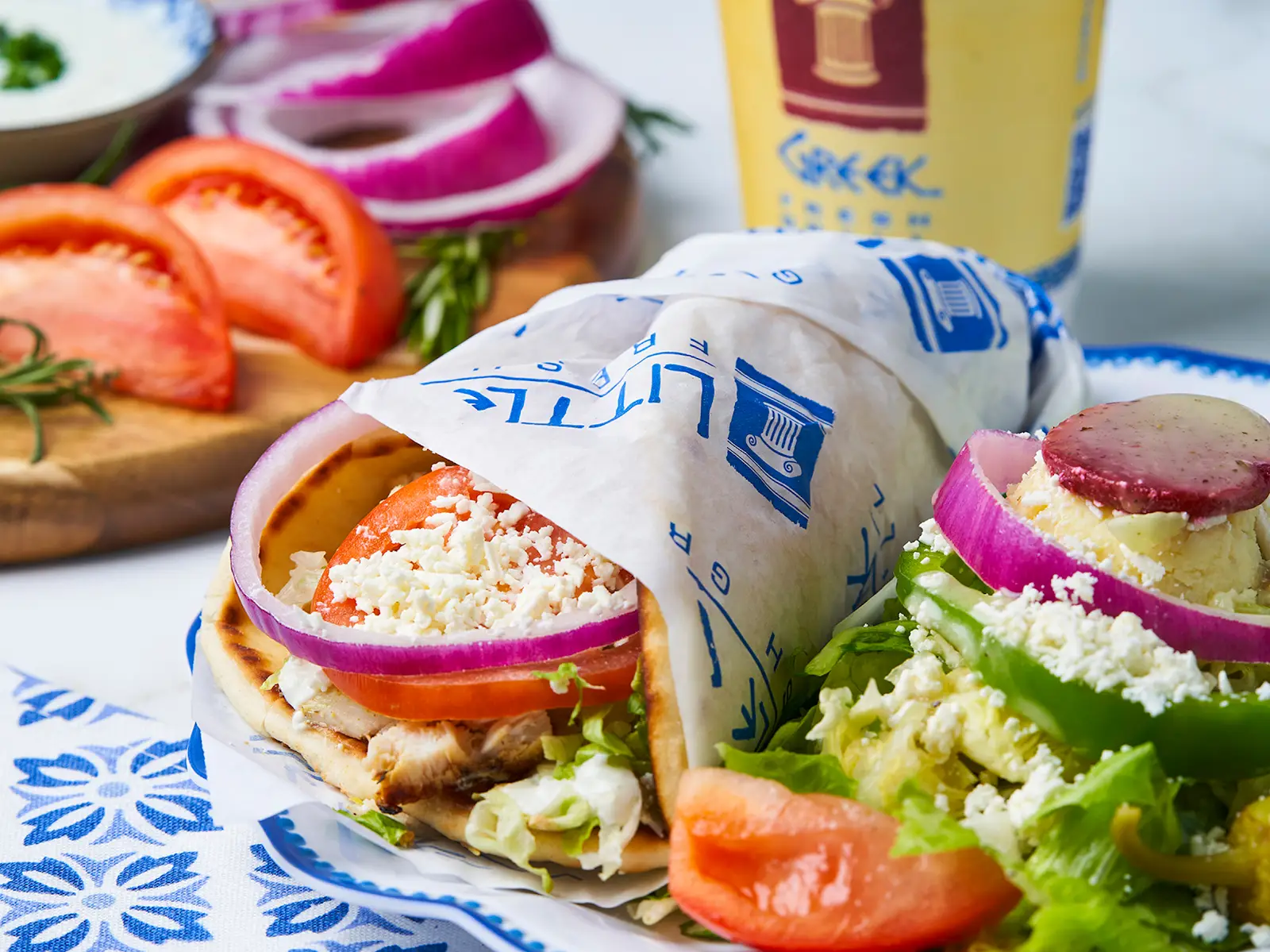 Little Greek Fresh Grill gyro in branded wrap paper with a Greek salad and branded drink cup
